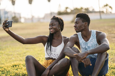 Paar nimmt Selfie durch Handy beim Sitzen auf Wiese während Sonnenuntergang - MPPF01890