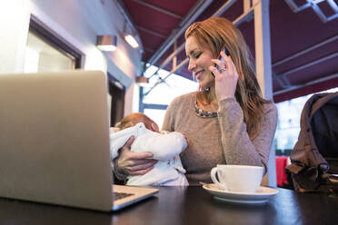 Mutter, die ihr Baby trägt, während sie in einem Café mit dem Handy telefoniert - WPEF04800