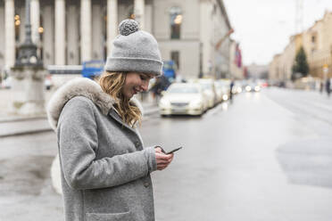 Lächelnde Frau mit Strickmütze, die auf einer Straße in der Stadt ein Mobiltelefon benutzt - WPEF04809