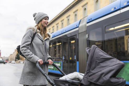 Lächelnde Frau, die einen Kinderwagen schiebt, während sie in der Stadt spazieren geht - WPEF04812