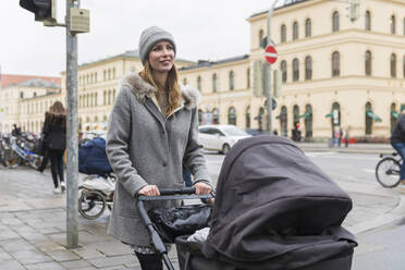 Lächelnde Frau schaut weg, während sie mit einem Kinderwagen in der Stadt spazieren geht - WPEF04814