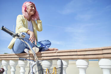 Cheerful young woman looking away while sitting on railing - KIJF03960