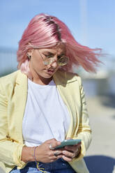 Junge Frau mit rosa Haaren benutzt ihr Smartphone am Strand - KIJF03964
