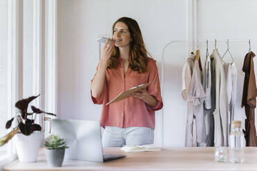 Female fashion designer with clipboard talking on mobile phone at home - AFVF08825