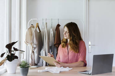 Weibliche Modedesignerin schreibt in ein Klemmbrett, während sie zu Hause im Büro sitzt - AFVF08831