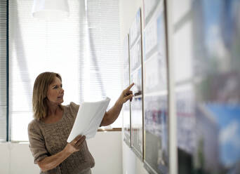 Businesswoman holding document while looking at charts on wall in office - AJOF01489