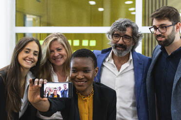 Lächelnde Geschäftsfrau nimmt Selfie durch Smartphone mit männlichen und weiblichen Kollegen im Büro - PNAF01842