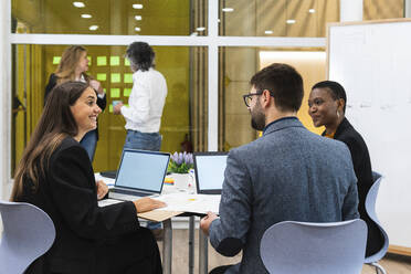 Smiling male and female entrepreneurs with laptop discussing during meeting with coworkers in background at office - PNAF01873