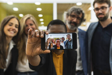 Lächelnde Geschäftsfrau nimmt Selfie durch Handy mit männlichen und weiblichen Mitarbeiter im Büro - PNAF01877