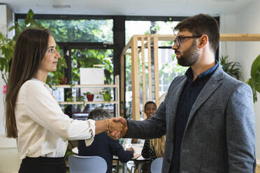 Young male and female entrepreneurs doing handshake with coworkers discussing in background at office - PNAF01894