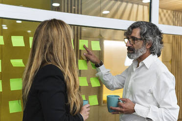 Senior businessman with coffee mug explaining business strategy to female coworker in office - PNAF01895