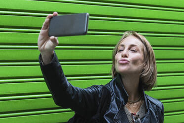 Frau schmiegt sich an, während sie ein Selfie mit ihrem Smartphone vor einem grünen Fensterladen macht - PNAF01897