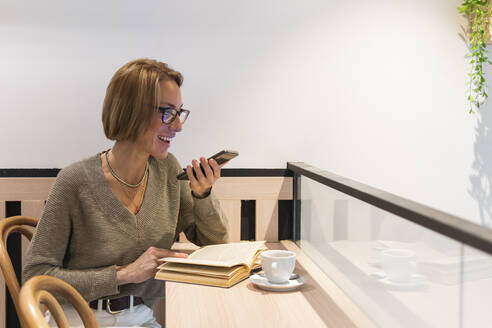 Lächelnde Frau mit Buch, die in einem Café mit ihrem Mobiltelefon spricht - PNAF01907