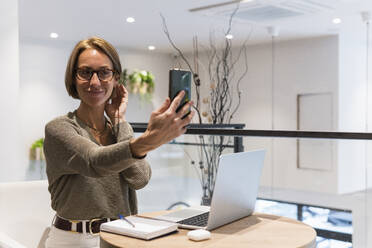 Lächelnde Frau mit Laptop, die ein Selfie mit ihrem Smartphone in einem Café macht - PNAF01910