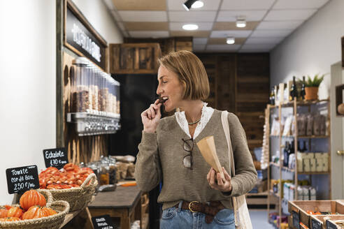 Blond woman eating food while looking at vegetables in supermarket - PNAF01920