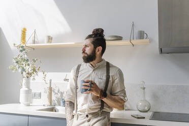 Nachdenklicher Mann mit Kaffeetasse, der wegschaut, während er zu Hause steht - EYAF01648