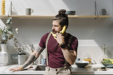 Mann benutzt Banane als Telefon, während er am Küchentisch steht - EYAF01658