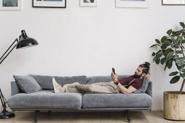 Man using mobile phone while lying on sofa in living room - EYAF01659