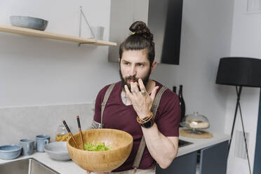 Bärtiger Mann, der in der Küche stehend Salat probiert - EYAF01660