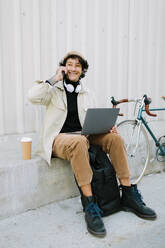 Lächelnder männlicher Freiberufler, der mit dem Handy telefoniert und einen Laptop benutzt - AGGF00114