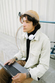 Thoughtful male freelancer wearing knit hat sitting with laptop while looking away - AGGF00116