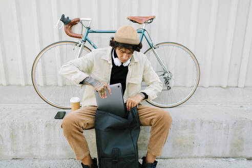 Männlicher Berufstätiger, der seinen Laptop in eine Tasche steckt, während er auf dem Gehweg sitzt - AGGF00117