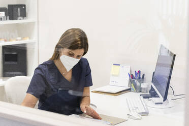 Zahnärztin mit Gesichtsschutzmaske bei der Arbeit am Schreibtisch in einer Klinik - PNAF01929