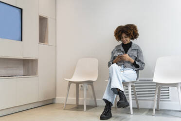 Smiling female patient using smart phone while sitting in waiting room at clinic - PNAF01957