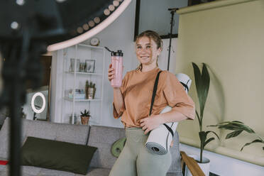 Smiling female influencer with exercise mat and smoothie vlogging at home - MFF08115