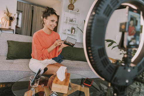 Female influencer showing make-up palette while doing tutorial at home - MFF08143