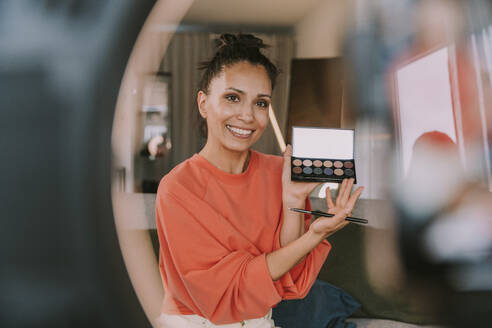 Smiling female influencer showing make-up palette while doing tutorial at home - MFF08145