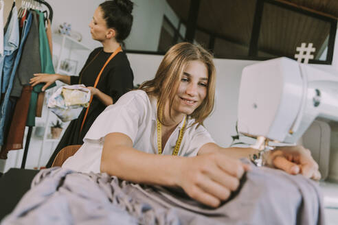Smiling teenage fashion designer sewing cloths with mature woman in background working at workshop - MFF08166