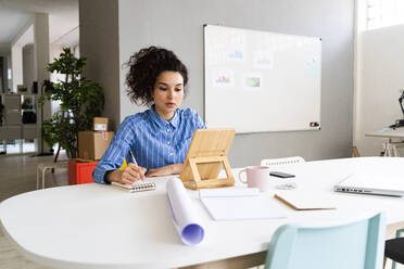 Young businesswoman writing in note pad while using digital tablet at office - GIOF12886