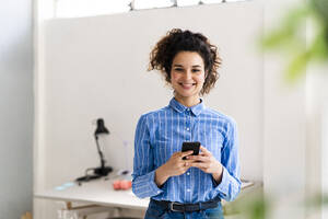 Lächelnde Geschäftsfrau, die ein Smartphone hält, während sie in einem kreativen Büro steht - GIOF12893