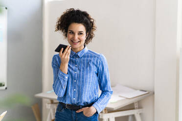 Lächelnde Geschäftsfrau mit Hand in der Tasche, die im Büro mit einem Mobiltelefon spricht - GIOF12895