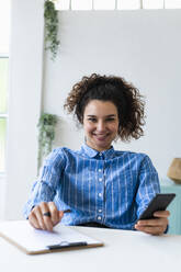 Lächelnde Geschäftsfrau mit Mobiltelefon am Schreibtisch im Büro sitzend - GIOF12910