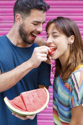 Glücklicher Mann, der einer Frau vor einer rosafarbenen Wellblechwand eine Wassermelone gibt - IFRF00825