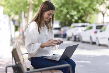 Lächelnde Frau, die ein Mobiltelefon benutzt, während sie mit einem Laptop auf einer Bank sitzt - JPF00422