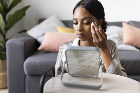 Young woman using tweezers on eyebrow while looking at mirror in living room - JPTF00822