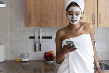 Junge Frau mit Handtuch, die in der Küche ein Mobiltelefon benutzt - JPTF00839