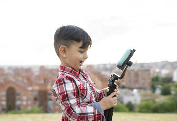 Boy smiling while holding tripod and mobile phone - JCCMF02854