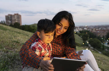 Smiling mother and son using digital tablet while sitting on hill - JCCMF02872