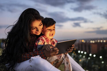 Happy son using digital tablet while sitting with mother during sunset - JCCMF02873