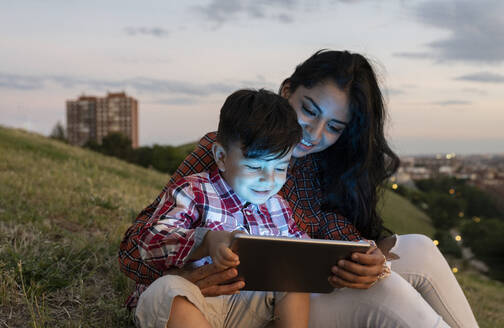 Frau und Junge benutzen digitales Tablet, während sie bei Sonnenuntergang auf einem Hügel sitzen - JCCMF02874