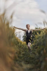 Bald transgender woman in black dress walking with arms outstretched in agricultural field - JPF00425