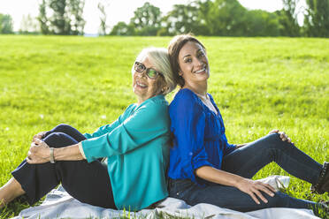 Senior woman sitting on picnic blanket with mid-adult woman at public park during sunny day - OIPF00879