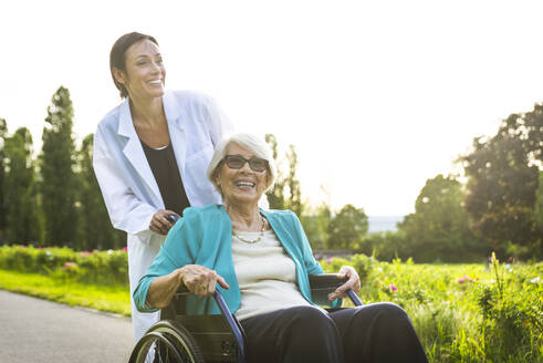 Lächelnde Pflegerin, die eine ältere Frau im Rollstuhl durch einen Park schiebt - OIPF00886