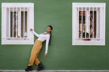 Young woman holding window grate by green wall outside house - TCEF01922