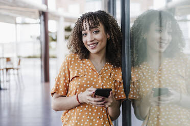 Lächelnde junge Frau mit Handy in der Hand, die sich an ein Fenster lehnt - EBBF03905