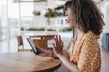 Lächelnde Frau grüßt während eines Videogesprächs über ein Mobiltelefon in einem Café - EBBF03910